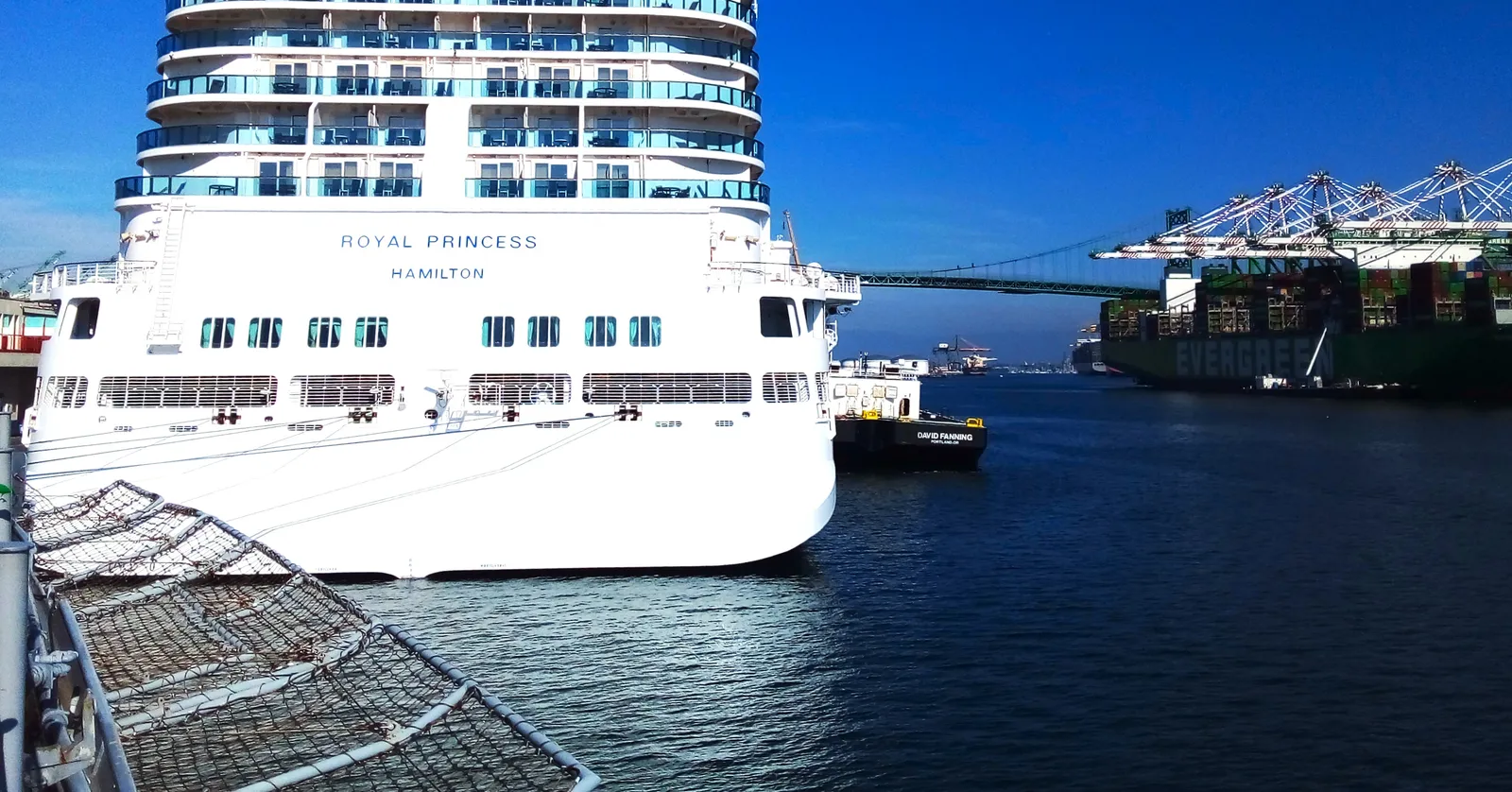 A white cruise ship tied up at the World Cruise Center in San Pedro, Los Angeles, with the green Vincent Thomas Bridge and container cranes across the main channel