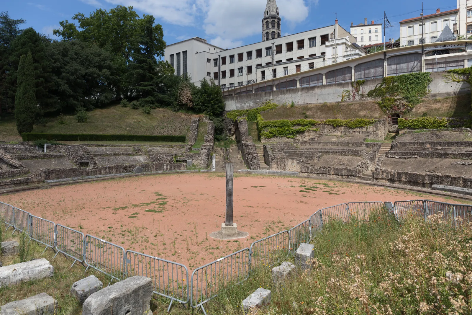 The remains of the Roman amphitheatre in Lyon, a sunken oval of sandy ground ringed by low stone terracing, with a plain memorial post standing at its centre and apartment blocks on the slope above