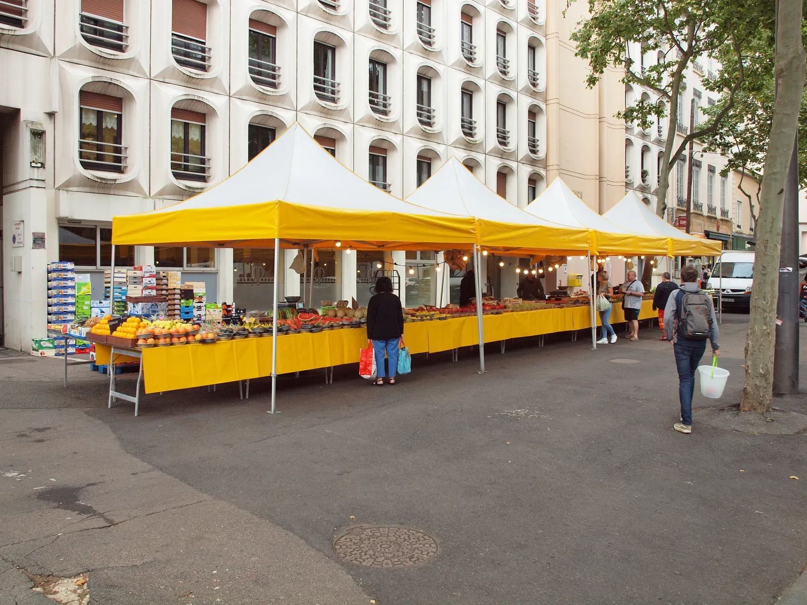 Market stalls with yellow canopies set up in a line along Boulevard de la Croix-Rousse in Lyon under plane trees, fruit and vegetables laid out on the tables and shoppers walking between them