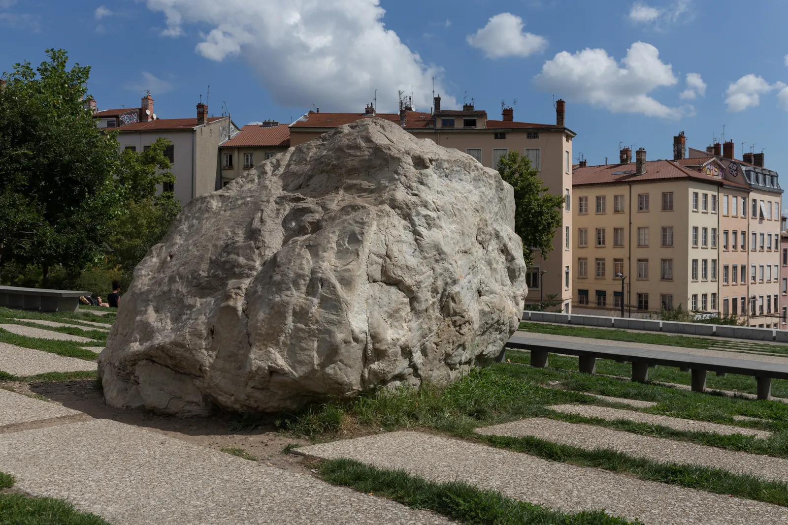 The Gros Caillou in Lyon, a pale boulder roughly the size of a small car sitting on grass beside a boulevard, with the coloured facades of Croix-Rousse apartment blocks behind it