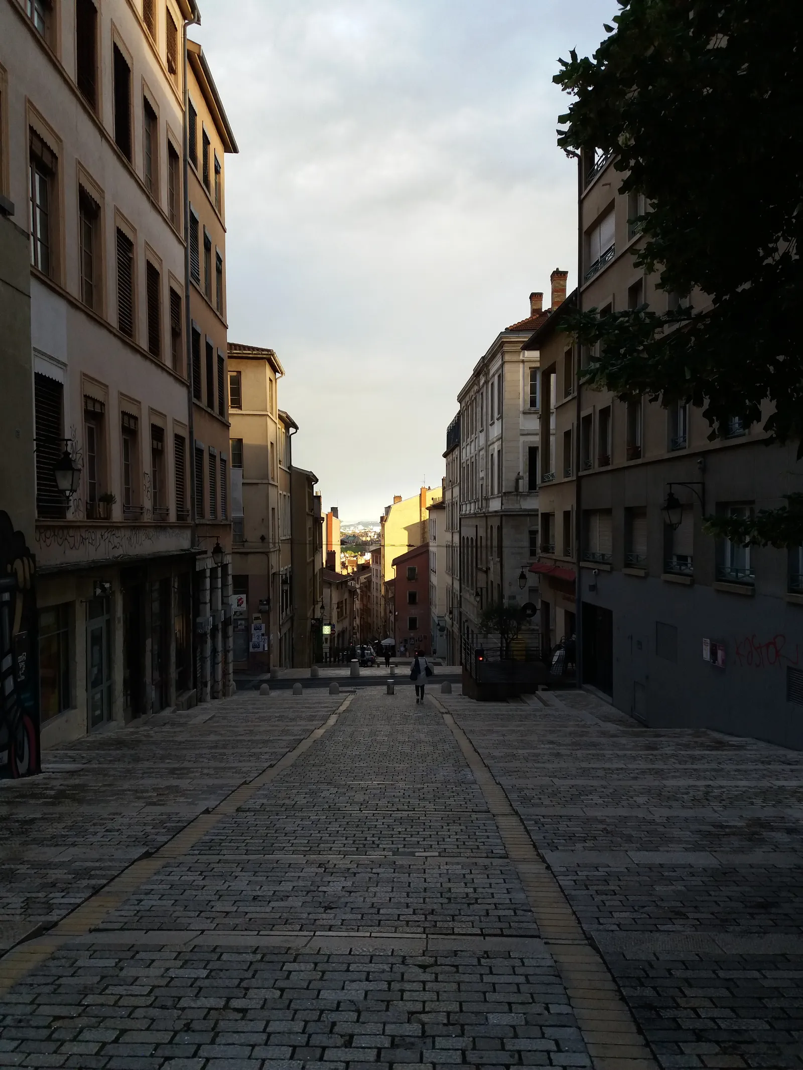 Looking down the Montée de la Grande Côte in Lyon at dusk, a wide cobbled street dropping in stages between tall shuttered buildings with the city visible far below at the bottom