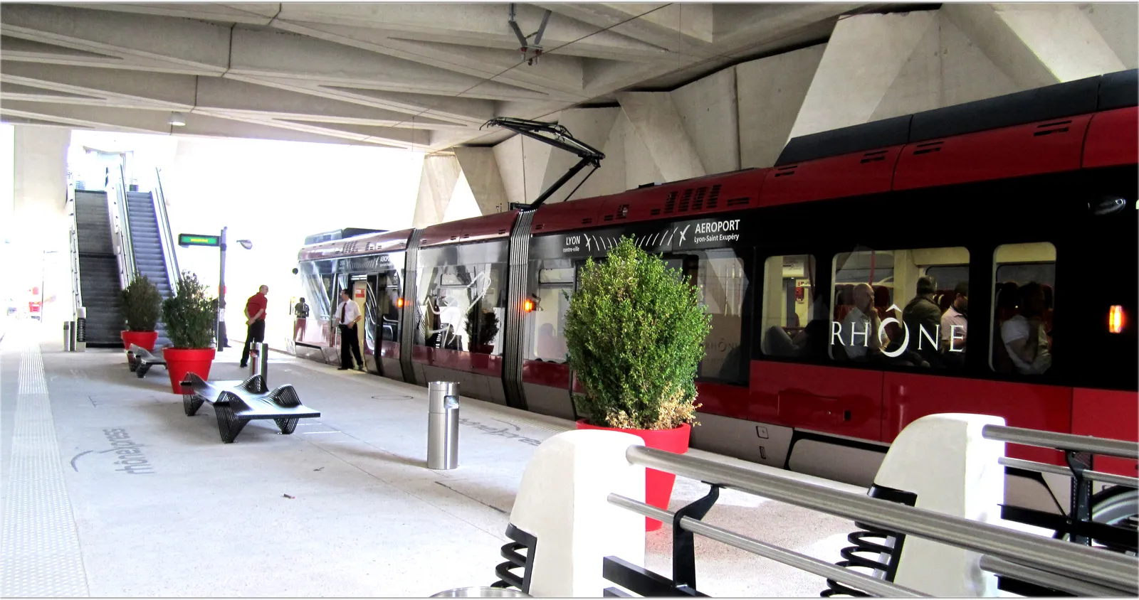 A red and grey airport tram standing at its platform under the concrete deck at Lyon-Saint Exupéry, passengers boarding through open doors with escalators up to the terminal behind