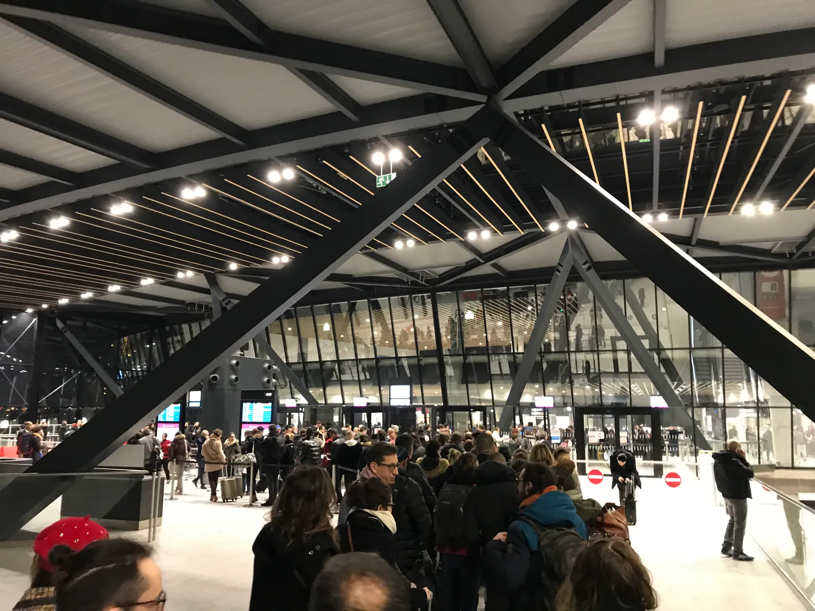 The departures hall of Terminal 1 at Lyon-Saint Exupéry Airport, heavy black steel bracing crossing under a slatted timber ceiling above a crowd of passengers waiting with bags