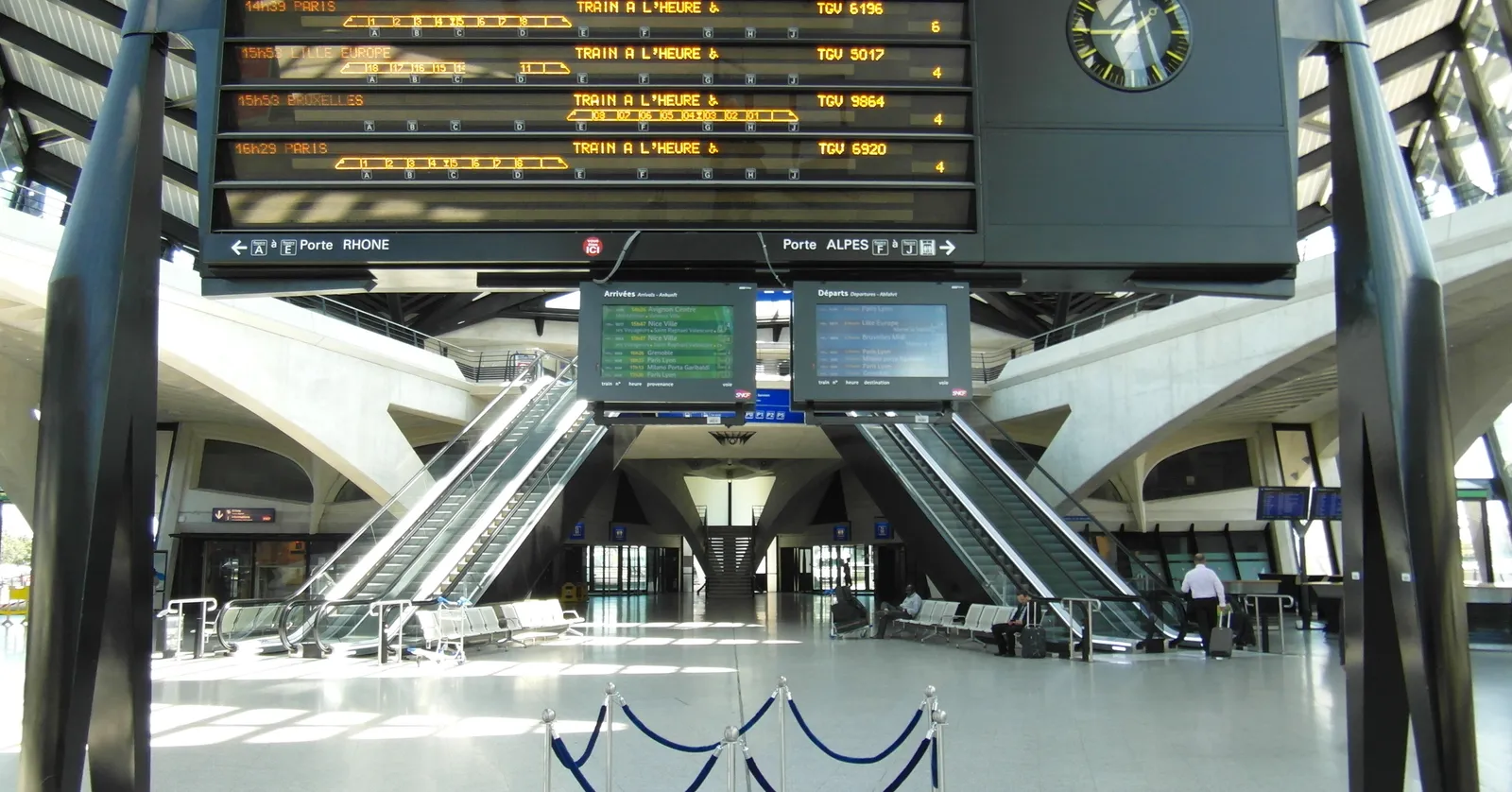 The main concourse of the railway station at Lyon-Saint Exupéry Airport, a ribbed white concrete vault over two banks of escalators with a large yellow departure board suspended above them