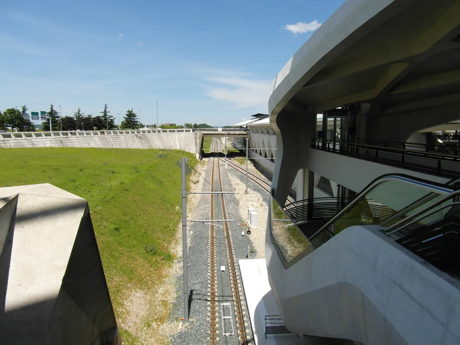 The outside of the railway station at Lyon-Saint Exupéry Airport, sculpted white concrete forms curving over the railway tracks, which run away into a covered tube under a blue sky