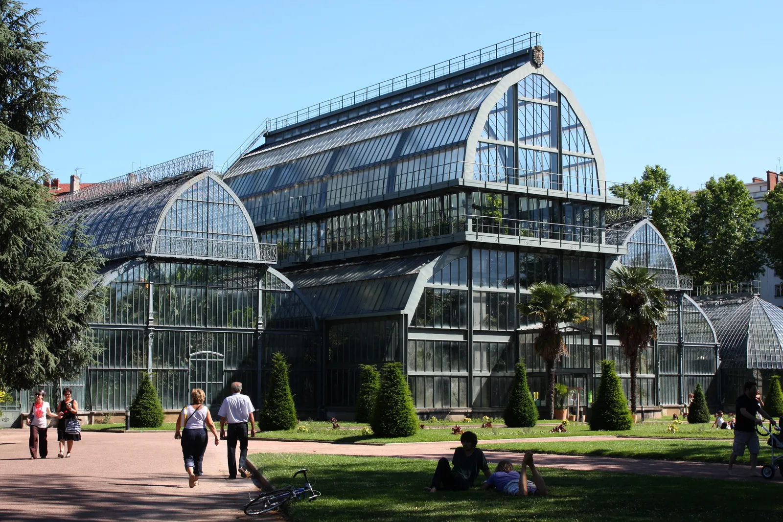 The large 19th-century glasshouse in the botanical garden at Parc de la Tête d'Or in Lyon, a tall iron and glass structure with curved gable ends, lawns and clipped shrubs in front