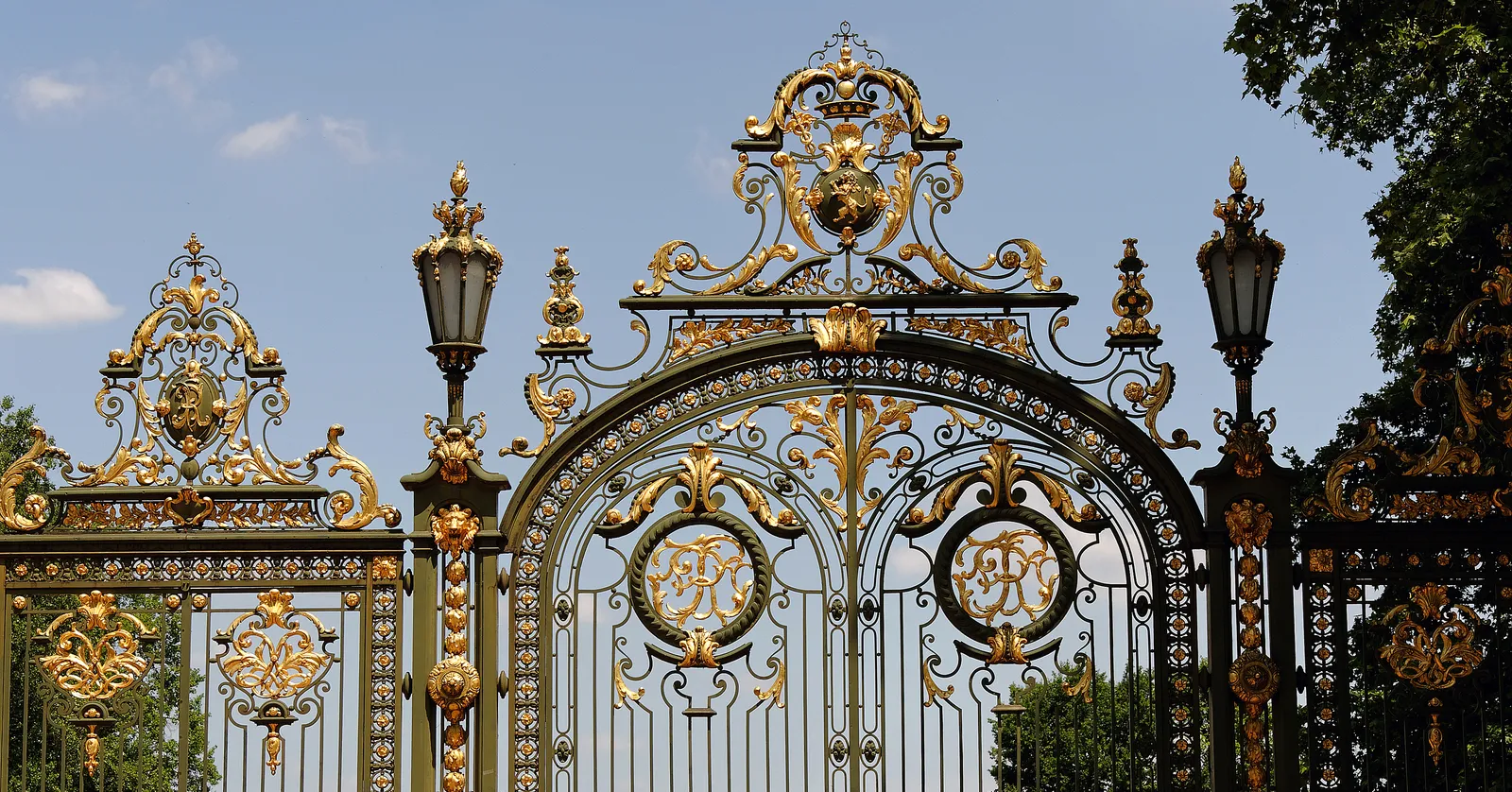 The main gate of Parc de la Tête d'Or in Lyon, tall wrought iron railings and scrollwork picked out in gold leaf with lanterns on the piers either side of the central arch