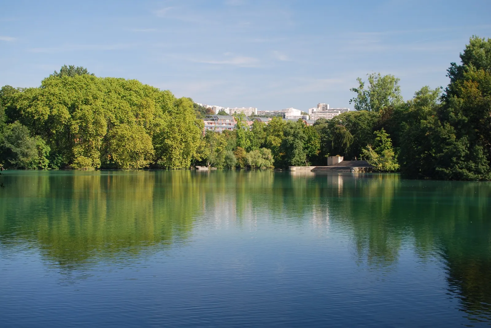 The lake in Parc de la Tête d'Or in Lyon on a still day, mature trees reflected across the water with a wooded island on the right and apartment blocks visible beyond the far bank