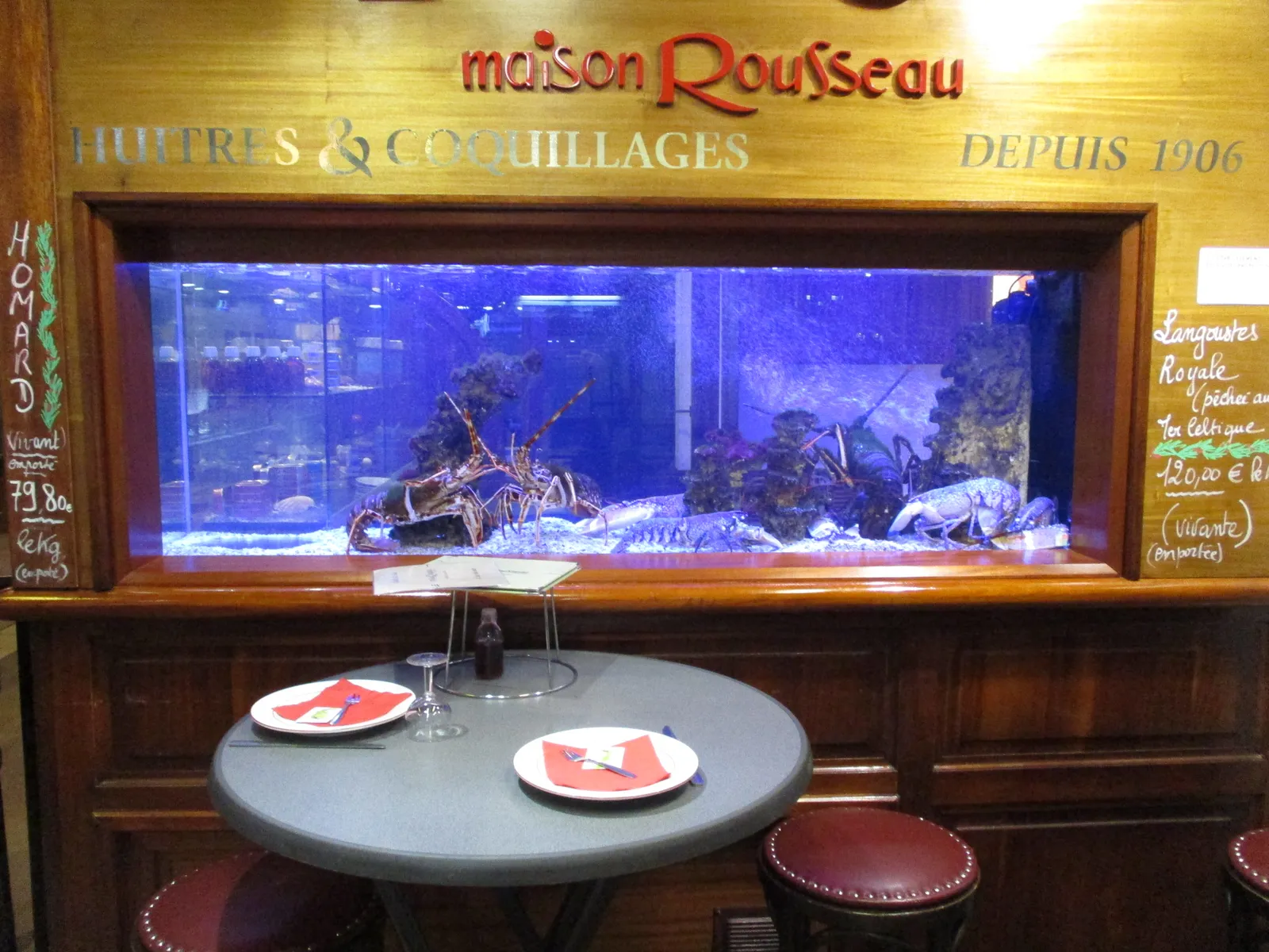 An oyster and shellfish stall inside Les Halles de Lyon Paul Bocuse, a lit tank of live lobsters set into a wooden counter with a small round table and two place settings in front of it