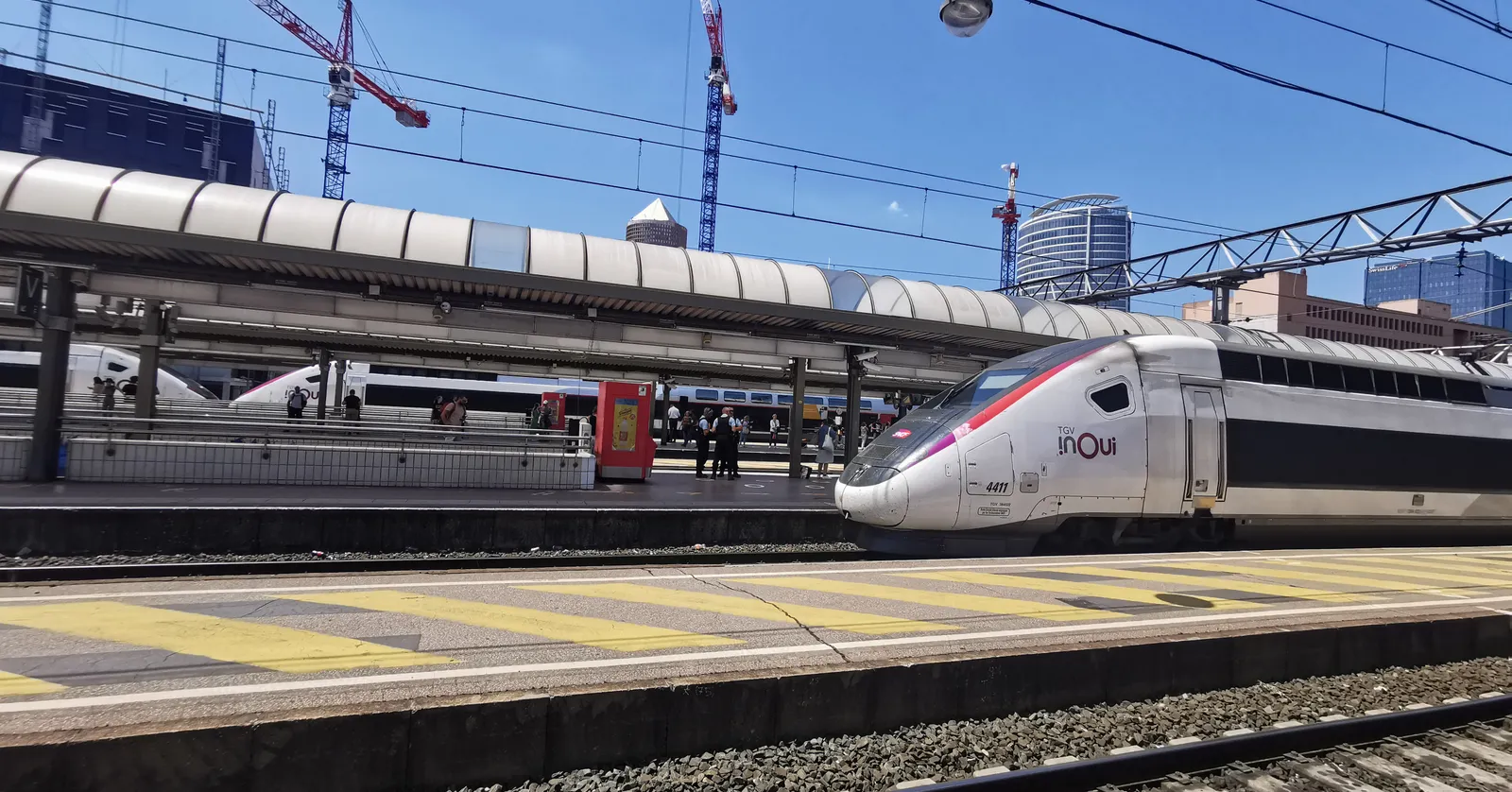 High-speed trains standing at the platforms of Lyon Part-Dieu station under a curved glass canopy, with the district's office towers and construction cranes behind