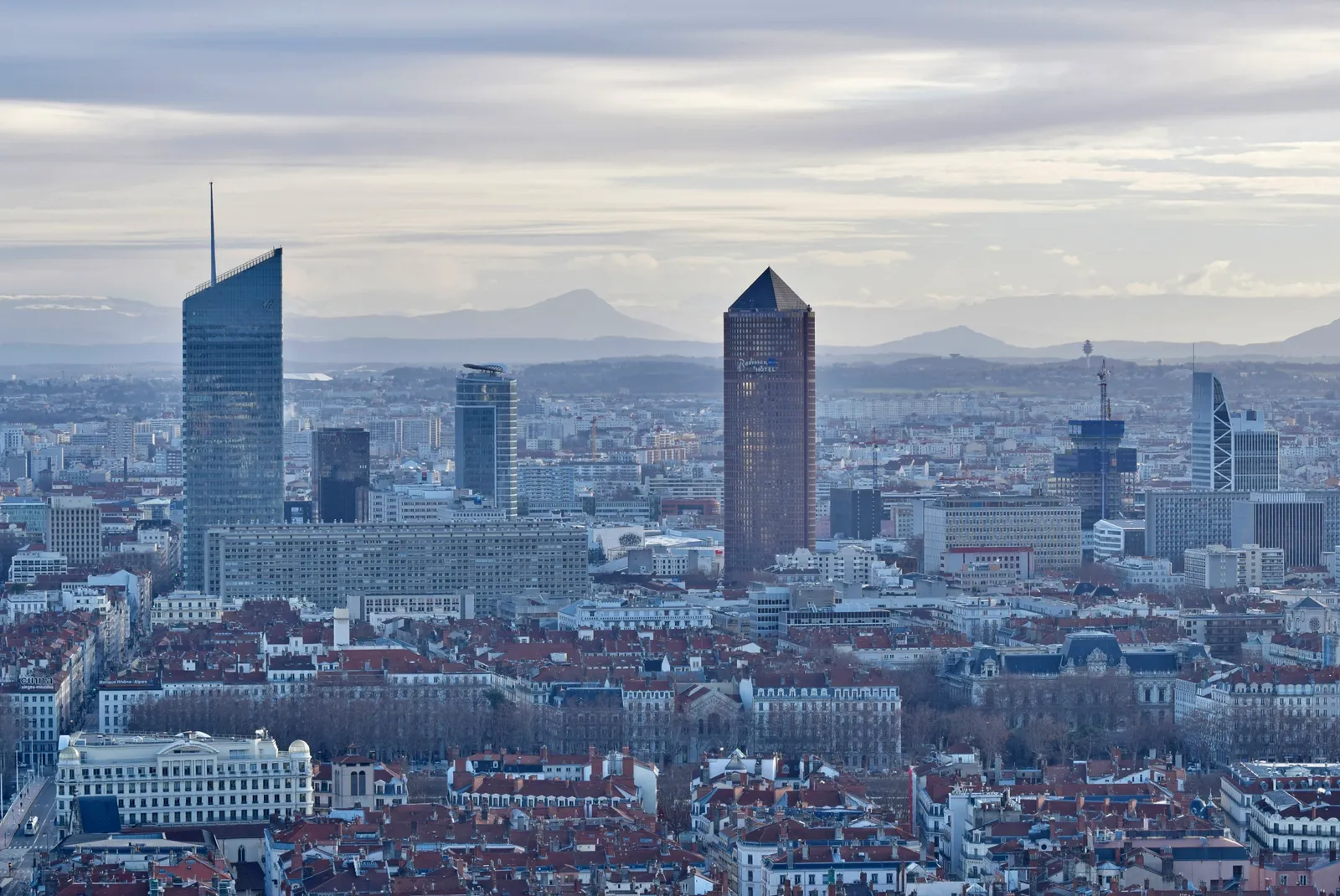 The Part-Dieu skyline in Lyon, a brown tower with a pointed pencil-shaped top standing among newer glass office towers above a spread of red-tiled rooftops