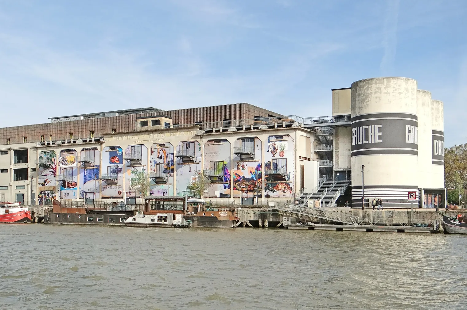 La Sucrière on the Saône quayside in Lyon, a long concrete former warehouse hung with large printed banners, with a white cylindrical silo tower at one end and barges moored in front