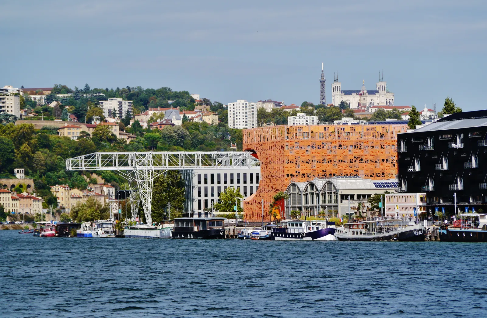 The Orange Cube on quai Rambaud in Lyon seen across the Saône, a bright orange perforated block beside an old dock crane and moored barges, with the Fourvière basilica on the hill behind