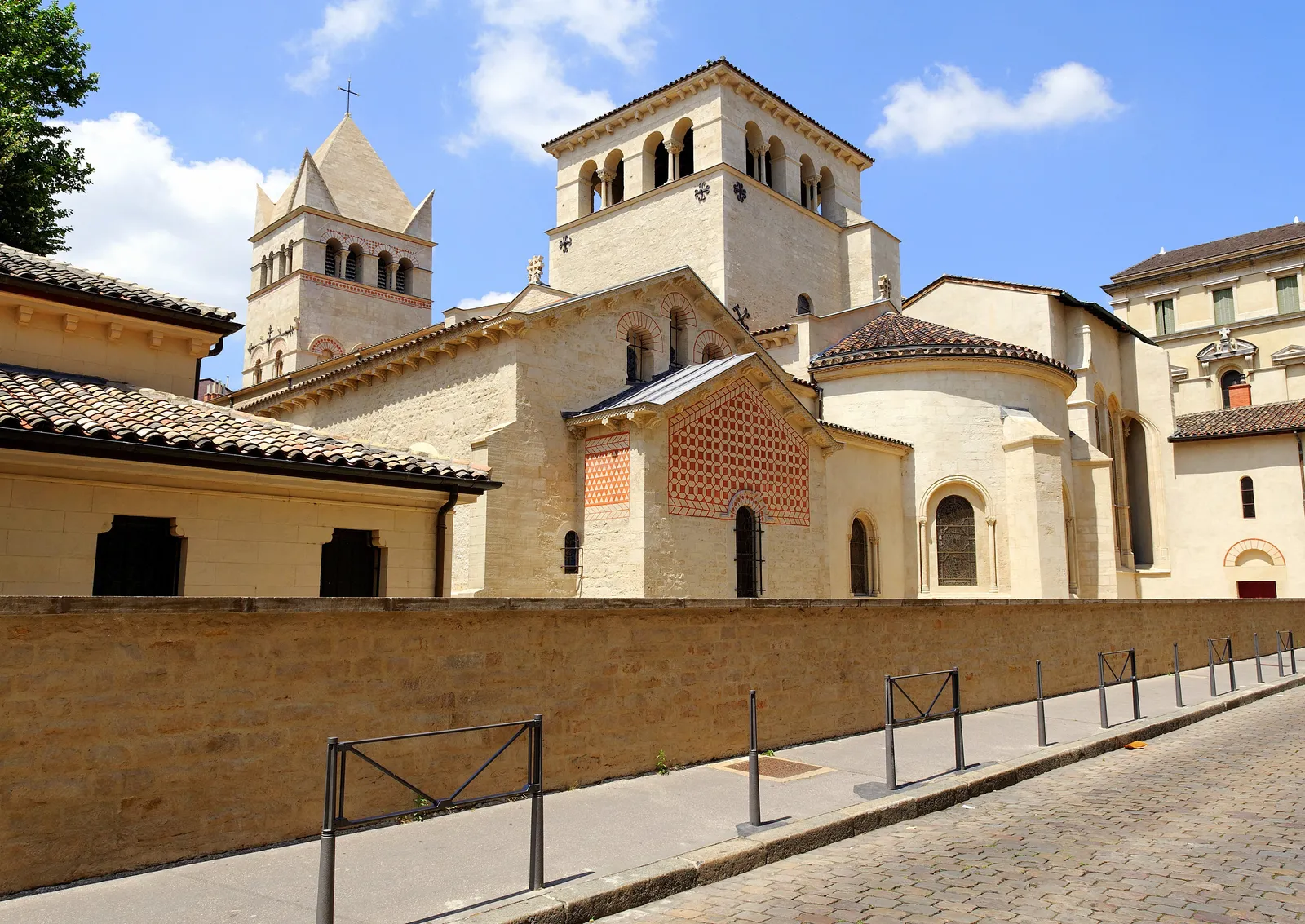 The Basilica of Saint-Martin d'Ainay in Lyon from the street, a low pale stone Romanesque church with a square bell tower and panels of red brick diamond patterning on its apse