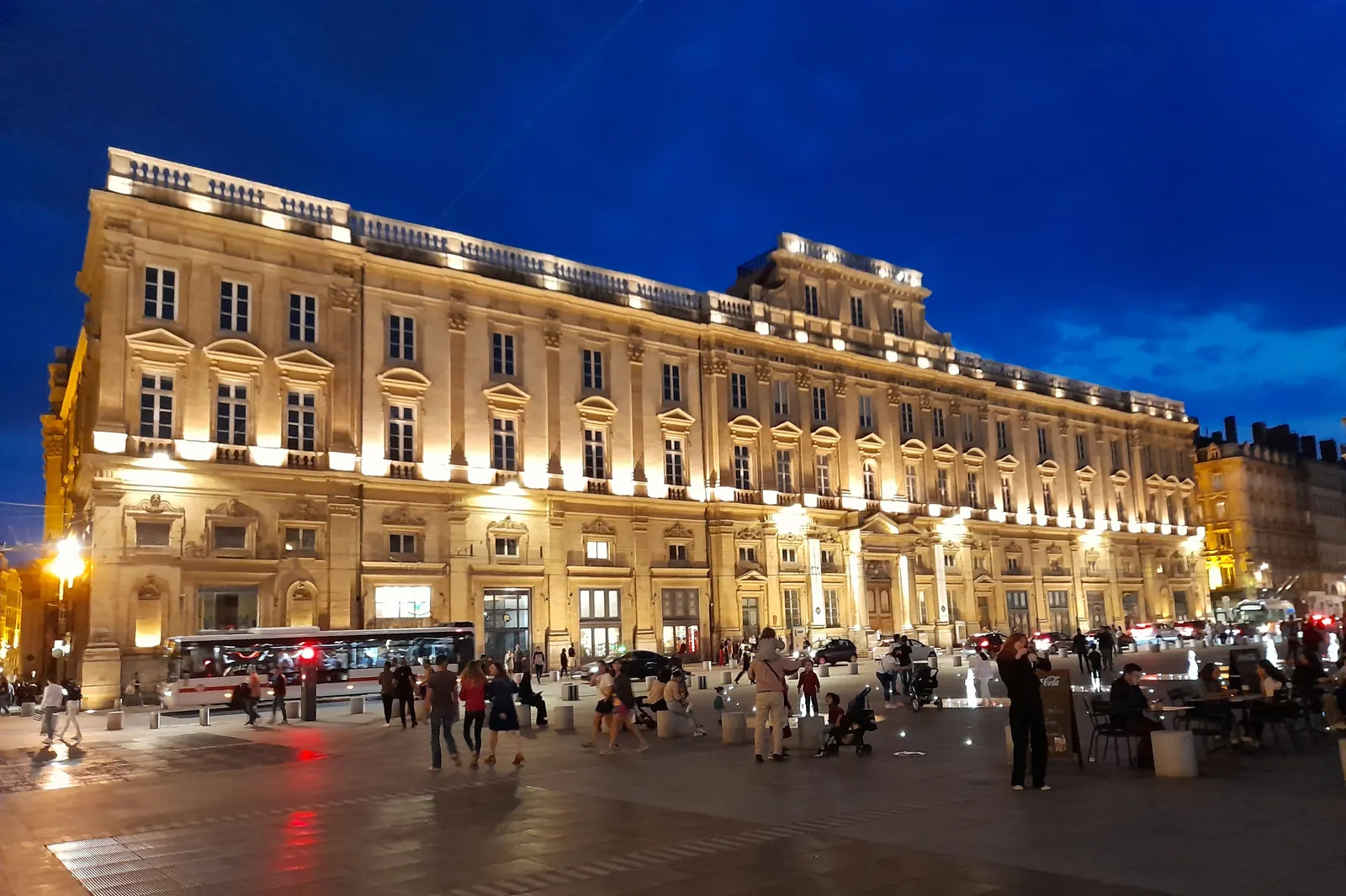 The Palais Saint-Pierre in Lyon lit up at night, a long 17th-century stone building housing the fine arts museum, with people crossing the square in front of it