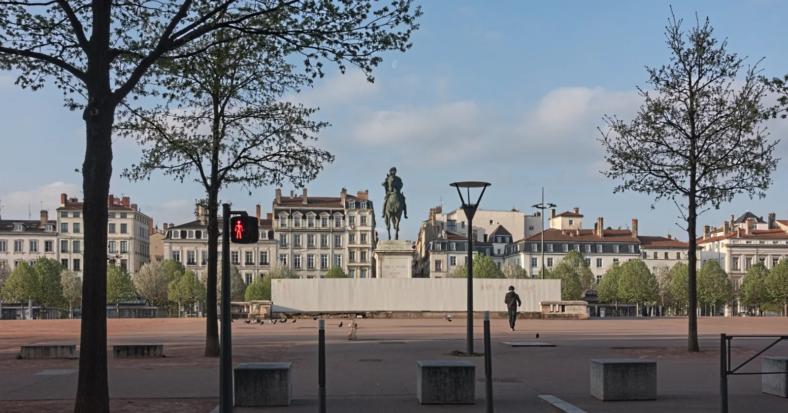 Place Bellecour in Lyon seen from its western side, the bronze equestrian statue of Louis XIV on a stone plinth in the middle of an open expanse of red gravel, with the square's uniform facades behind