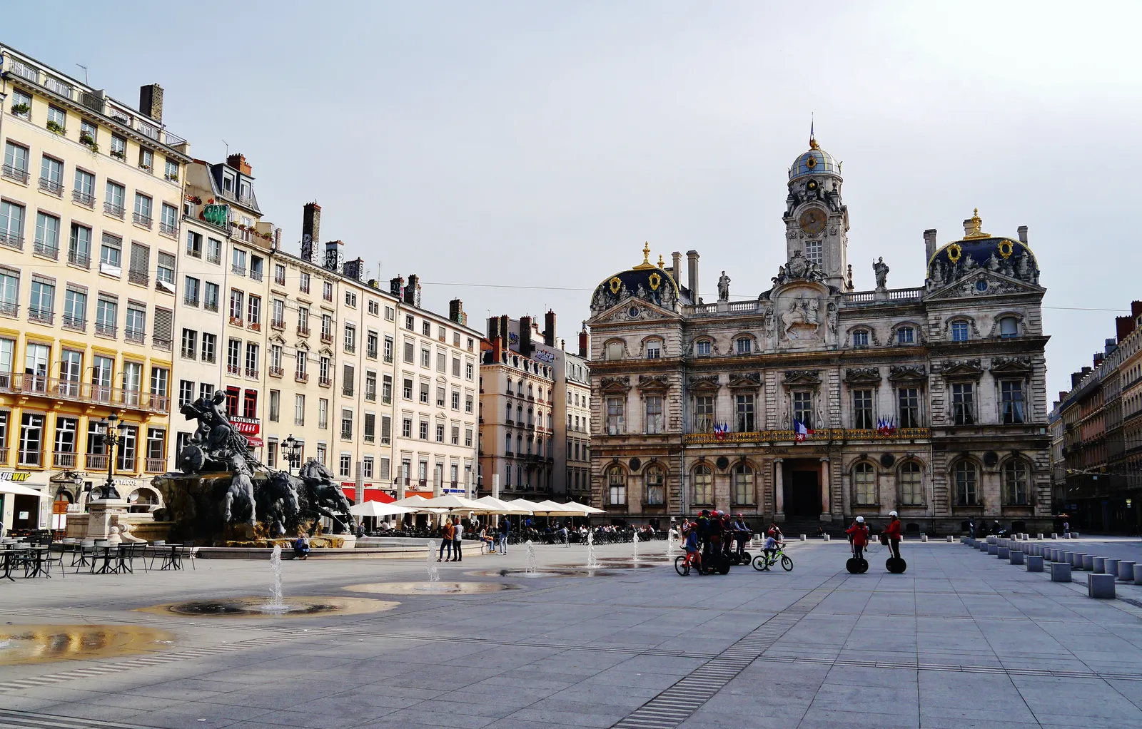 Place des Terreaux in Lyon, the ornate town hall filling the eastern side of the square, the dark bronze Bartholdi fountain with its four horses on the left and low ground fountains across the paving