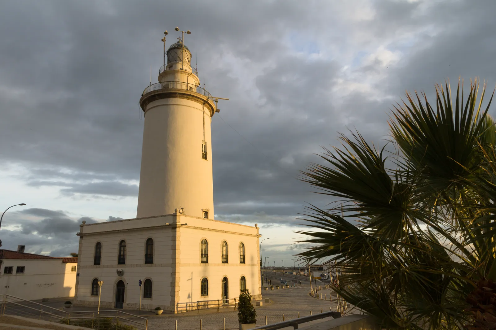 La Farola lighthouse in Málaga at dusk, a tall white cylindrical tower at the end of the harbor pier