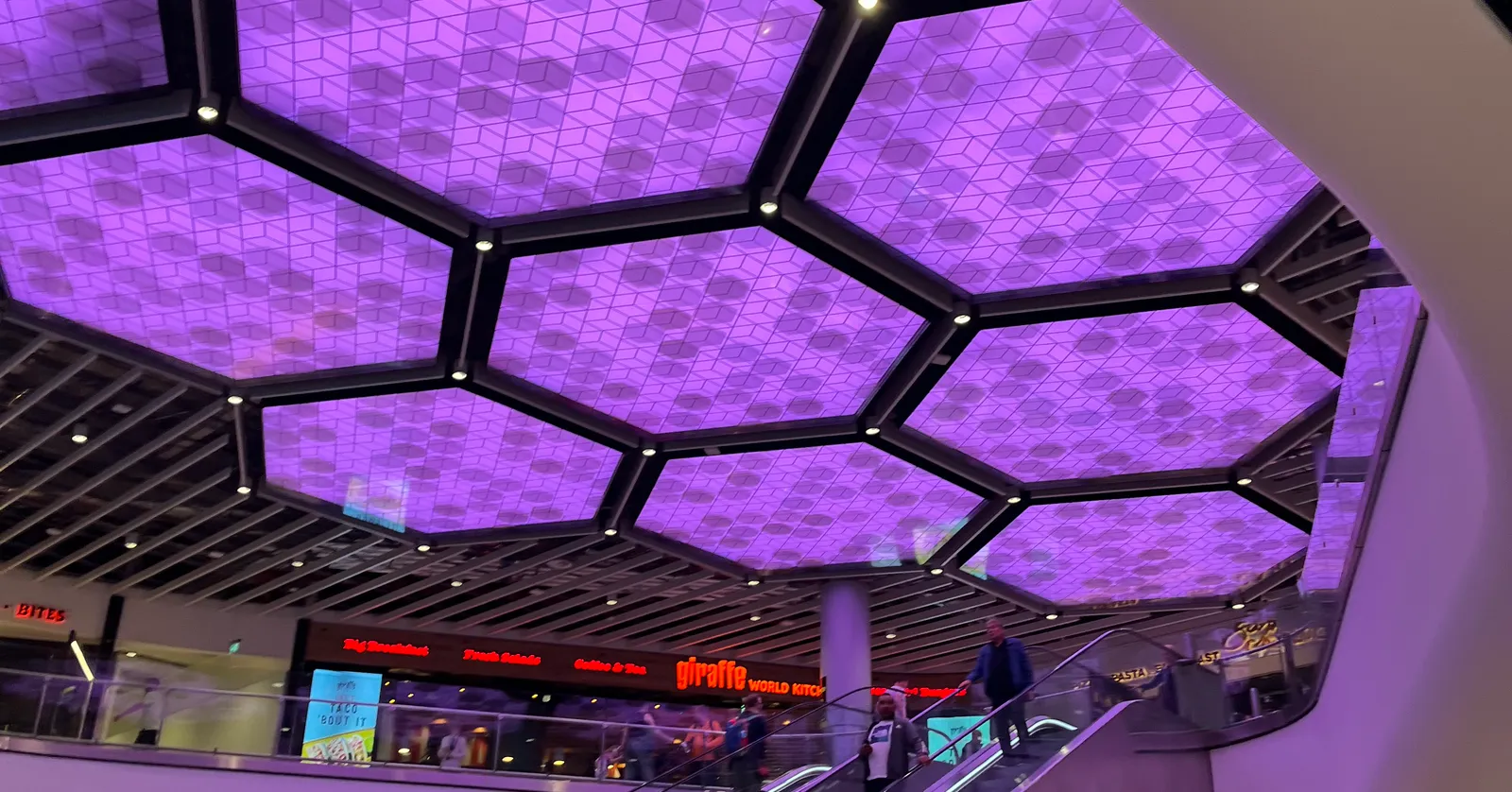 A hexagonal LED ceiling feature above an escalator in Manchester Airport's rebuilt Terminal 2, with restaurant signage including Giraffe World Kitchen visible below