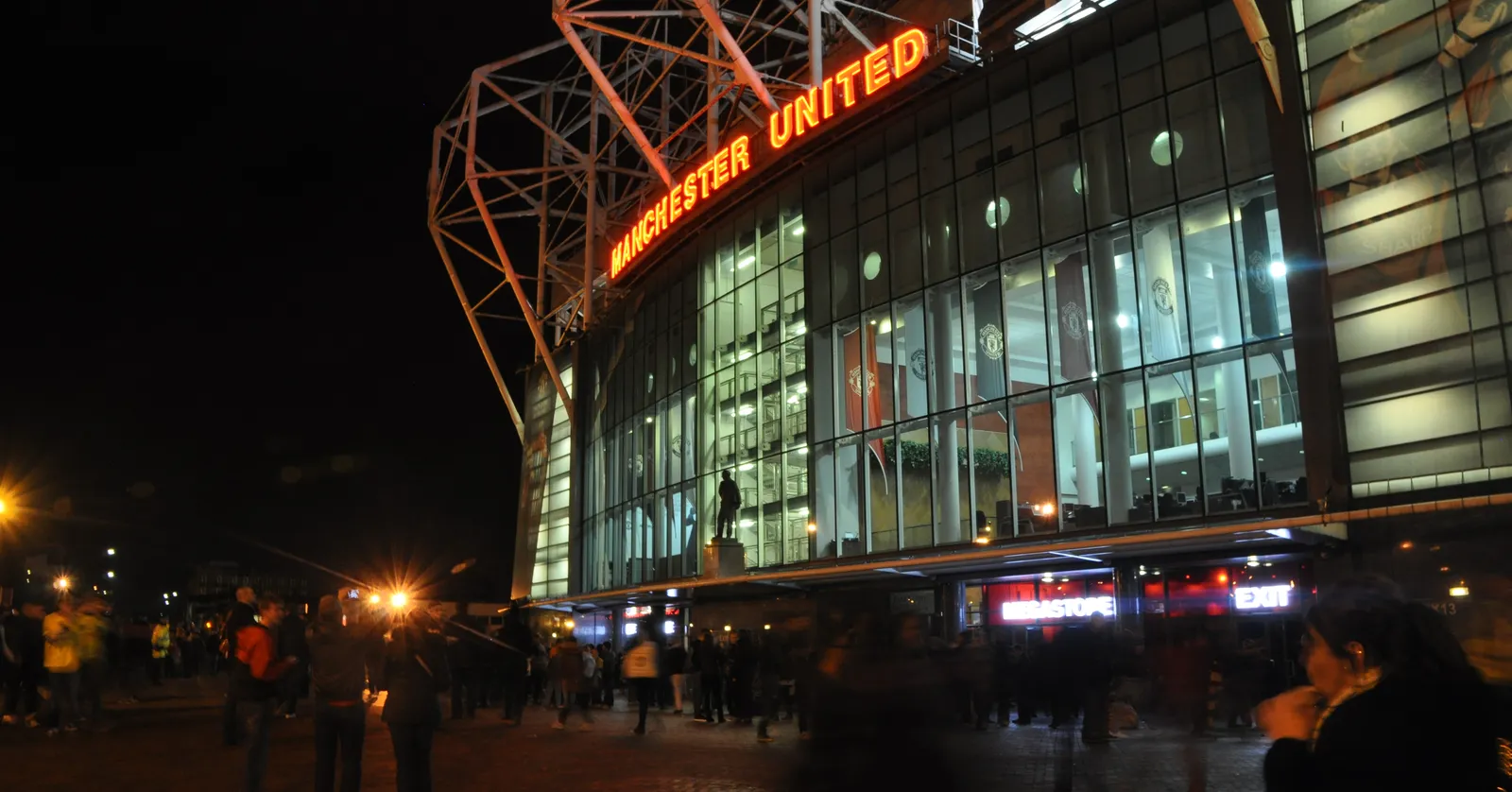Old Trafford stadium lit up at night, with the illuminated red MANCHESTER UNITED sign across the glass forecourt entrance