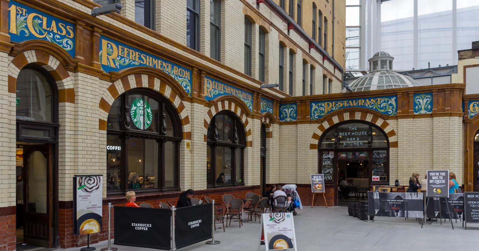 The restored Edwardian arcade inside Manchester Victoria station, with ornate tiled shopfronts reading '1st Class Refreshment Room' under a glass roof