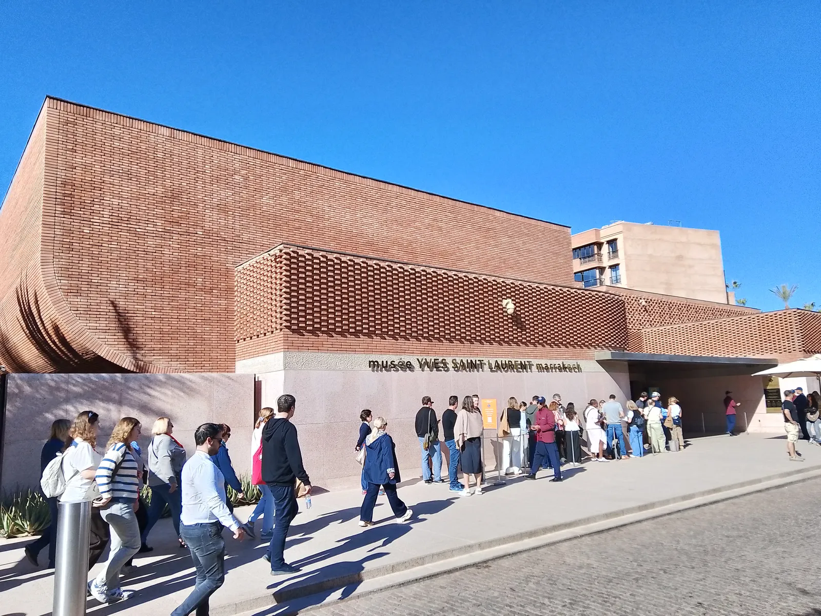The Musée Yves Saint Laurent on Rue Yves Saint Laurent in Marrakech, a low building of terracotta brick with a band of lattice brickwork above the entrance, visitors queuing along the pavement under a clear blue sky