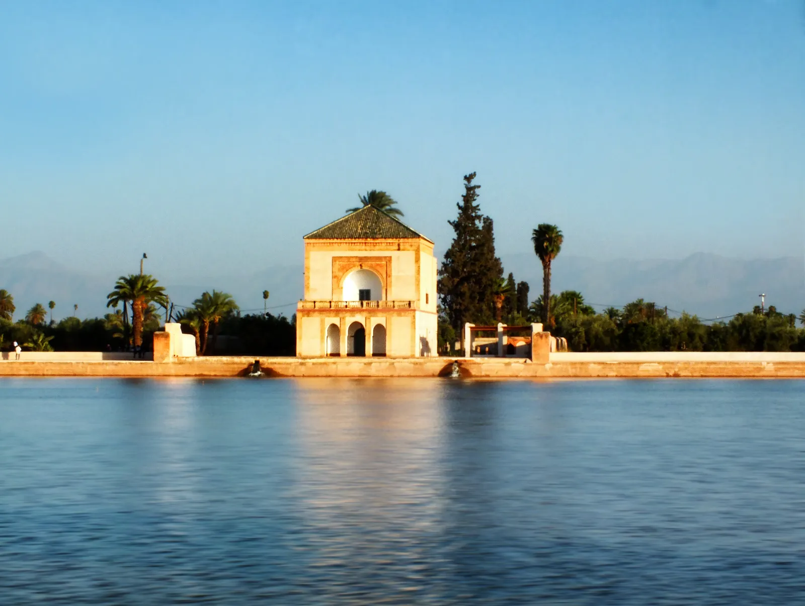 The Menara pavilion in Marrakech, a square two-storey building with a green tiled pyramid roof standing at the edge of a wide still basin, cypresses and palms beside it and the grey outline of the Atlas mountains on the horizon