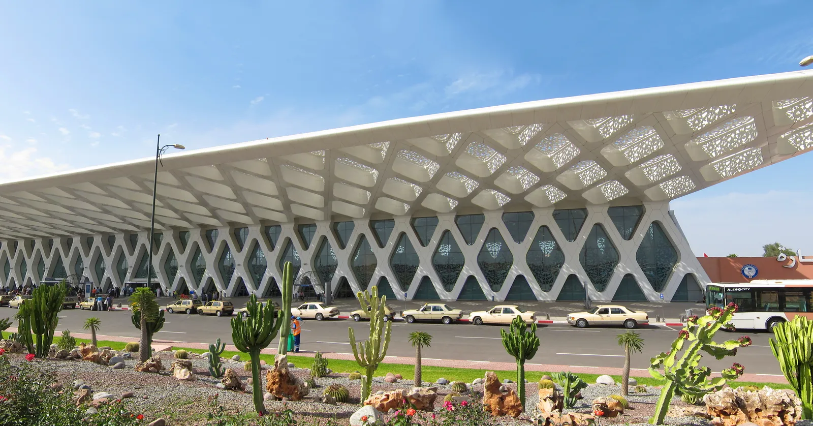 Terminal 1 at Marrakesh Menara Airport, a long white roof resting on a lattice of diamond-shaped concrete frames with glass between them, cacti and palms planted along the forecourt and a bus parked at the kerb