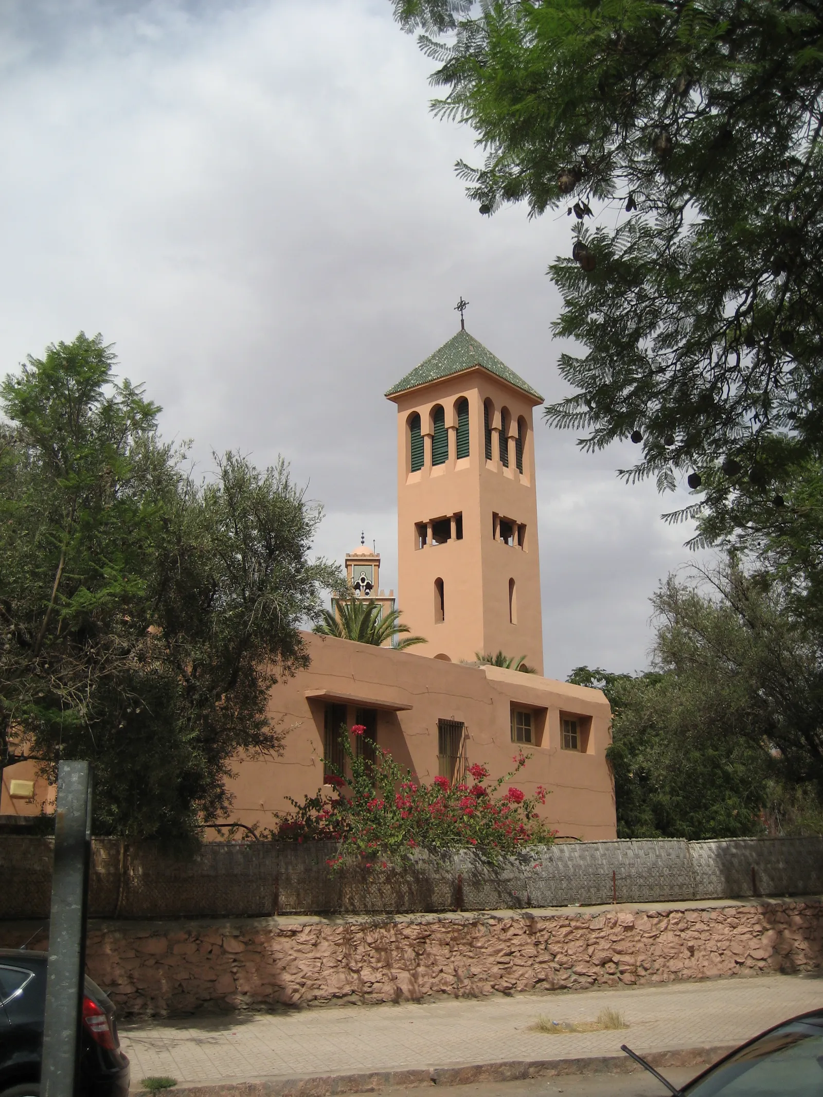 The Église des Saints-Martyrs in Gueliz, Marrakech, a salmon-pink church with a square bell tower under a green tiled roof and a cross, trees and bougainvillea along its wall and a mosque minaret visible behind