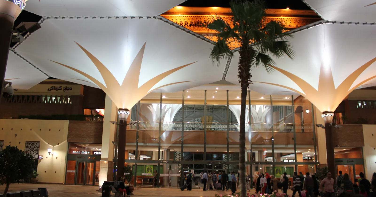 The forecourt of Marrakech railway station at night, white fabric canopies spreading from tall columns like leaves, a palm tree in a flower bed in the middle and travelers with wheeled bags walking toward the lit glass entrance