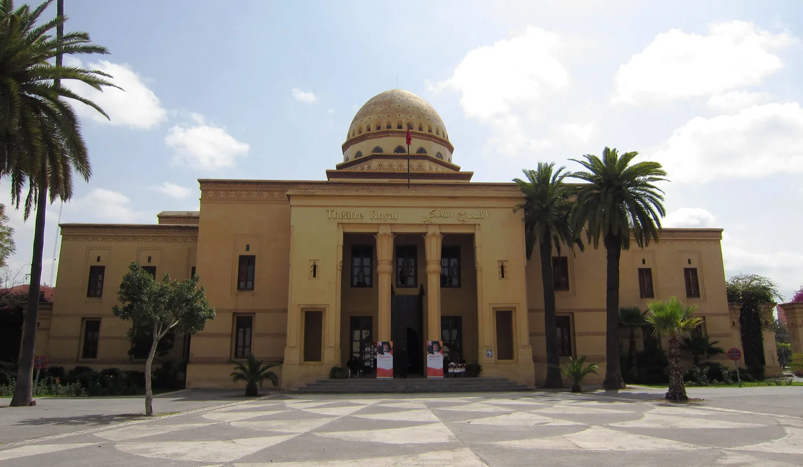The Théâtre Royal in Marrakech, a symmetrical sand-coloured building with a colonnaded entrance under a large ribbed dome, palm trees on either side and a patterned paved plaza in front