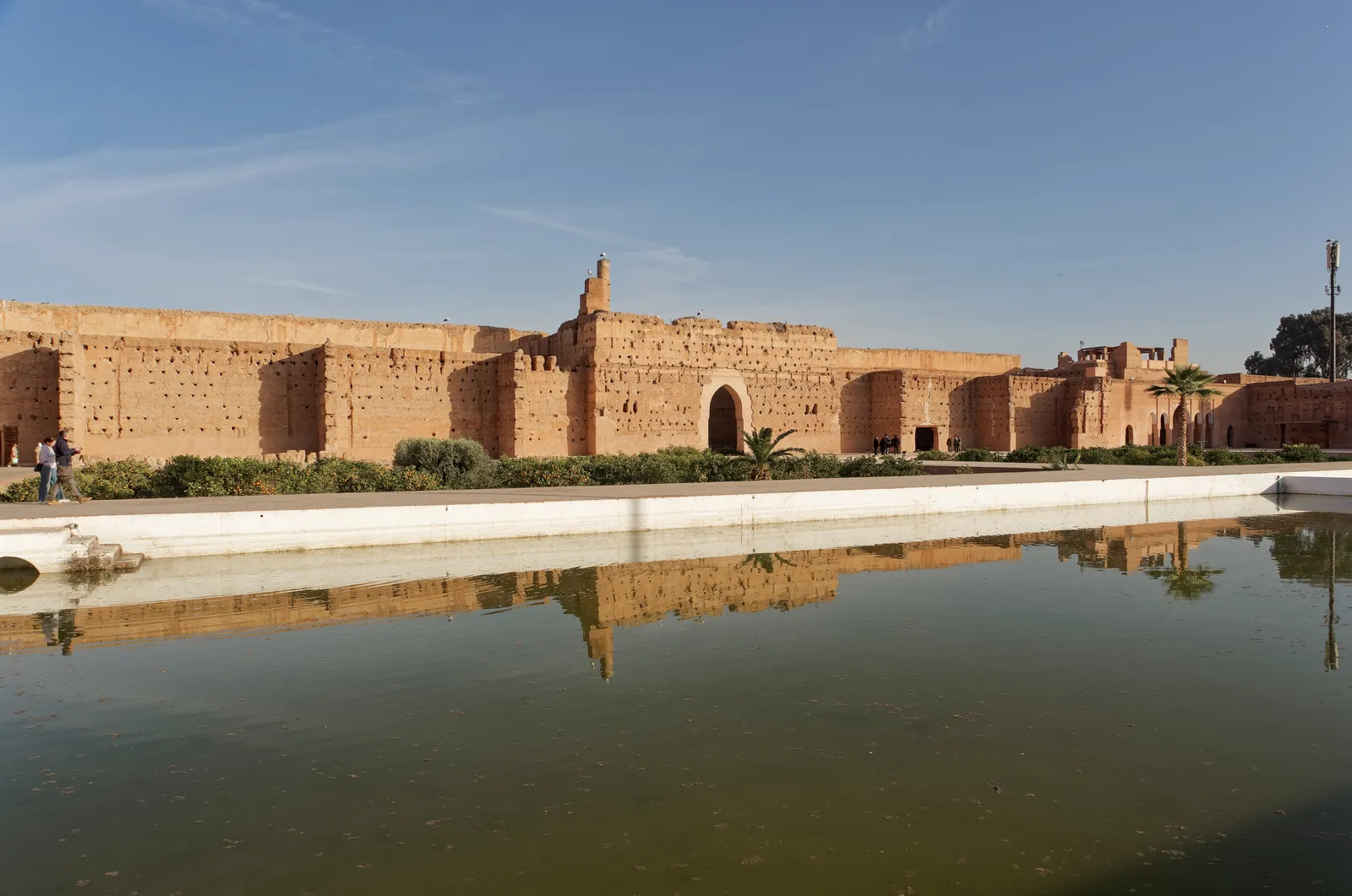 The courtyard of the El Badi Palace in Marrakech, a long rectangular pool of still green water in the foreground reflecting the ruined pink earthen walls and a gateway tower on the far side