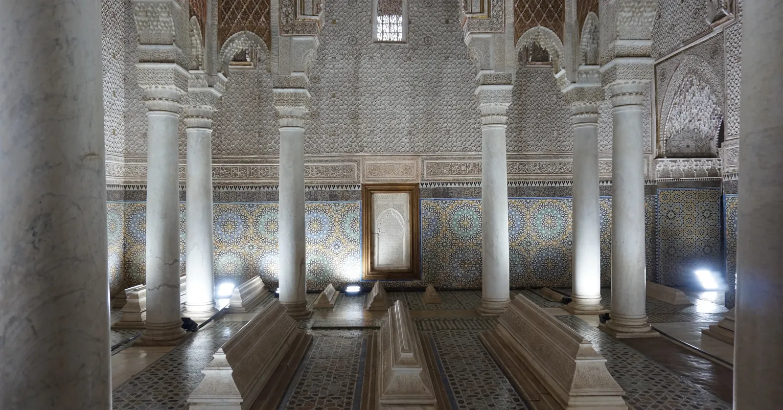 The Hall of Twelve Columns in Marrakech's Saadian Tombs, white marble columns in rows around low tiered marble tombs, walls of zellige tilework and carved stucco lit from below