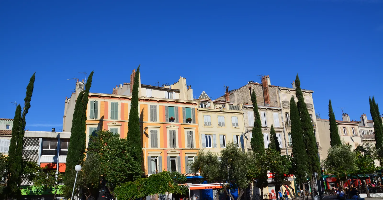 The painted ochre and cream facades along Cours Julien in Marseille, cypresses and olive trees rising in front of them above shopfronts and café awnings