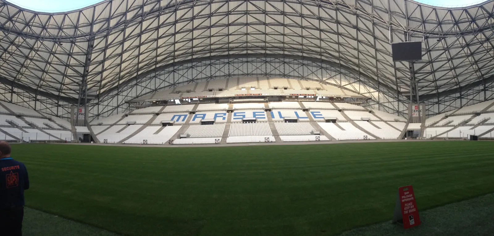The empty bowl of the Orange Vélodrome in Marseille seen from pitch level, MARSEILLE picked out in white seats across the stand under the stadium's curved white lattice roof