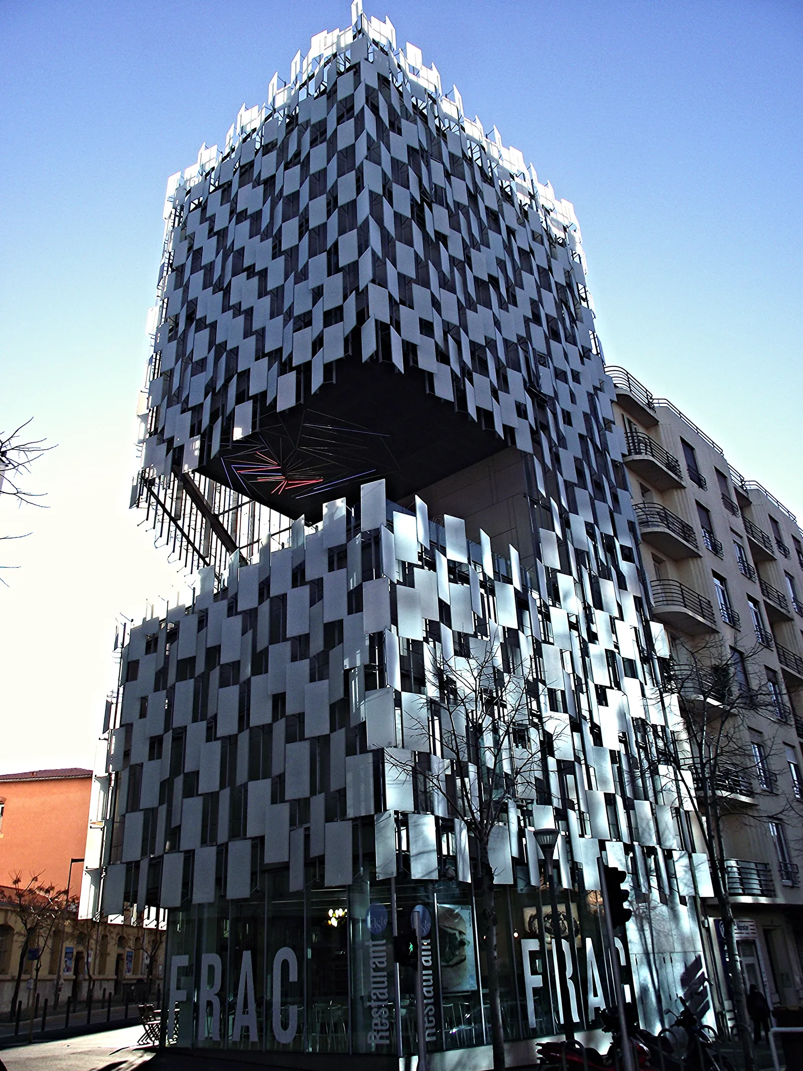 The Frac Sud building in Marseille, a dark cube clad in hundreds of tilted white and grey glass panels, cantilevered over a glazed ground floor lettered FRAC