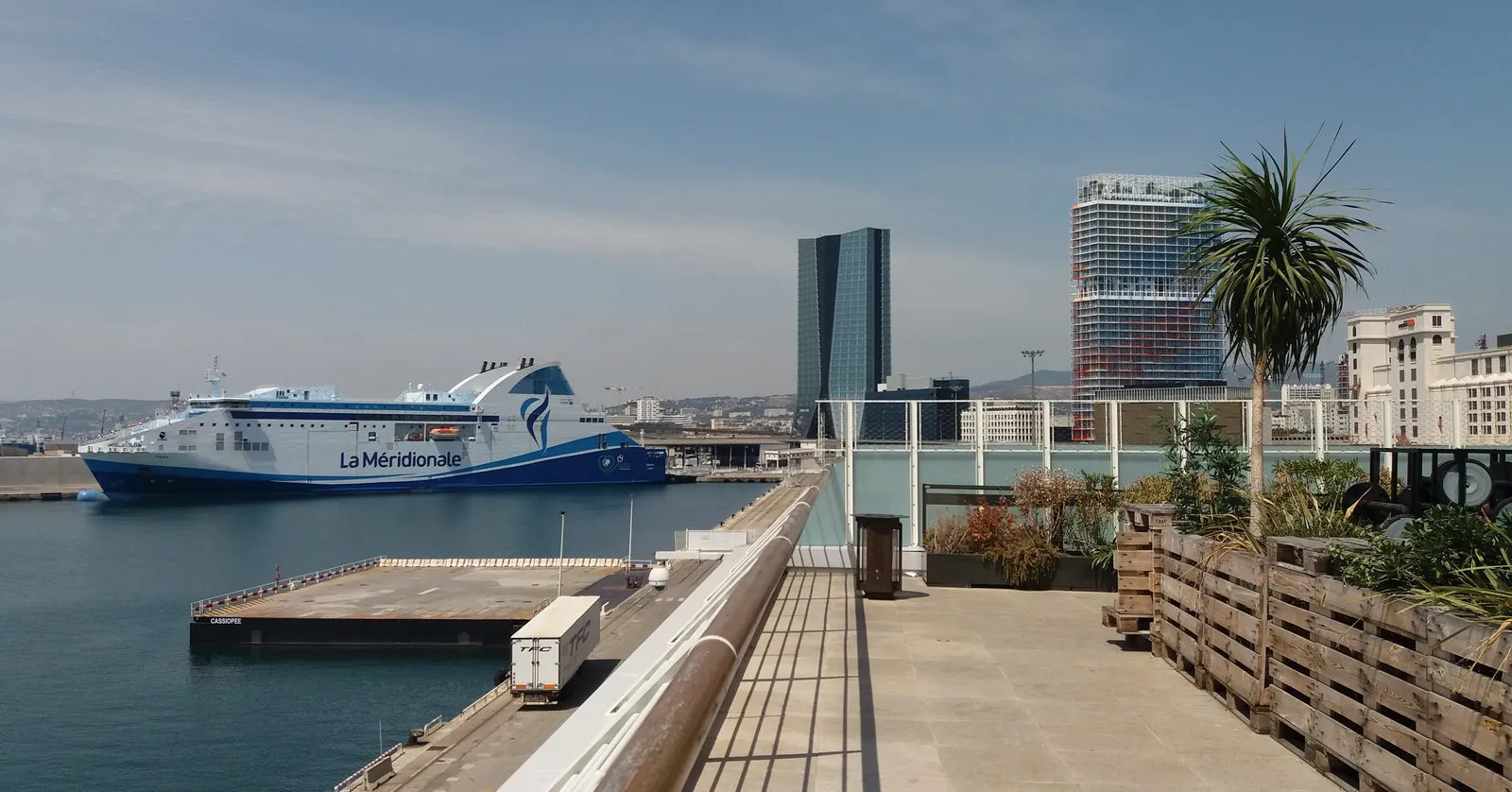 The open sea terrace at Les Terrasses du Port in Marseille, its railing running along the quay with a blue and white passenger ferry moored at the berth below and the harbour-front towers behind