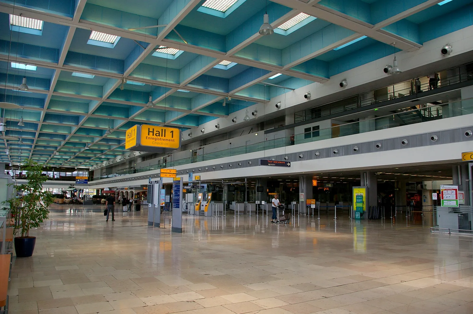 The concourse of Hall 1 at Marseille Provence Airport, a wide polished floor under a deep turquoise coffered concrete ceiling punched with square skylights