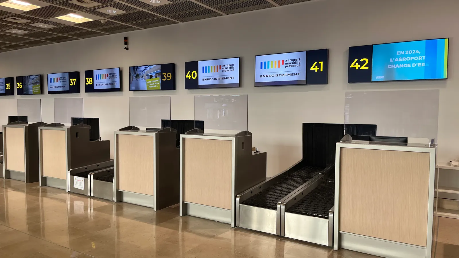 A row of numbered check-in desks in Terminal 1 at Marseille Provence Airport, bag belts set into pale wood counters under screens showing each desk number