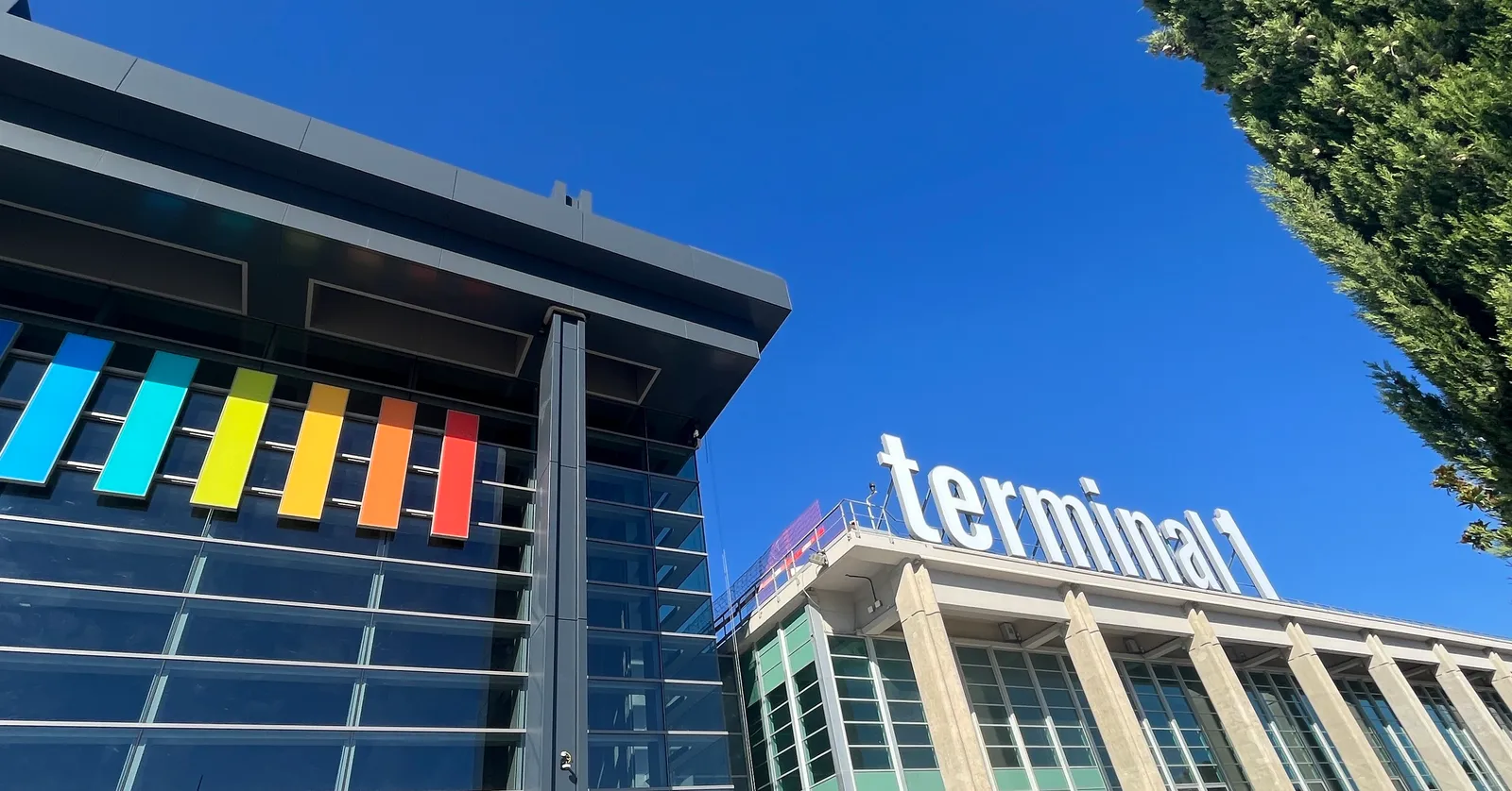 The frontage of Terminal 1 at Marseille Provence Airport, white lettering reading terminal 1 standing on the roof of a concrete-finned building beside a glass block hung with coloured panels