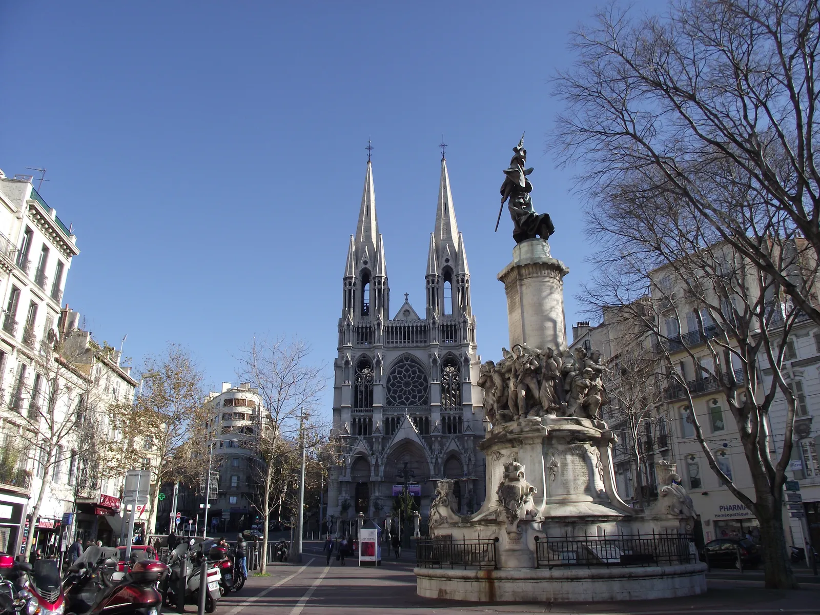 The neo-Gothic front of Les Réformés church in Marseille with its two stone spires, seen from the tree-lined top of the Canebière beside a carved public fountain