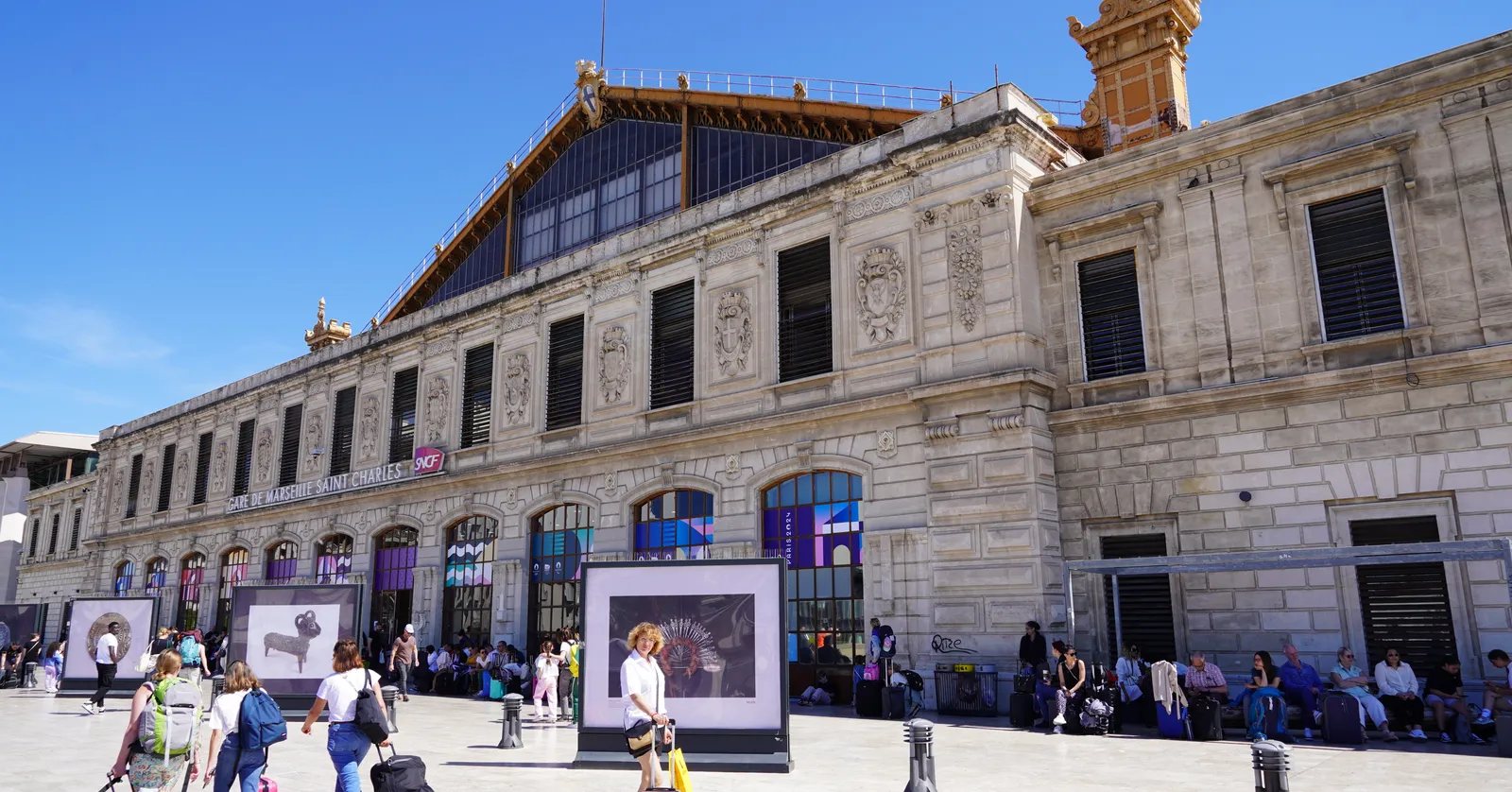 The stone facade of Marseille Saint-Charles station seen from its forecourt, lettered Gare de Marseille Saint Charles above the entrance doors, with travellers wheeling suitcases across the paving