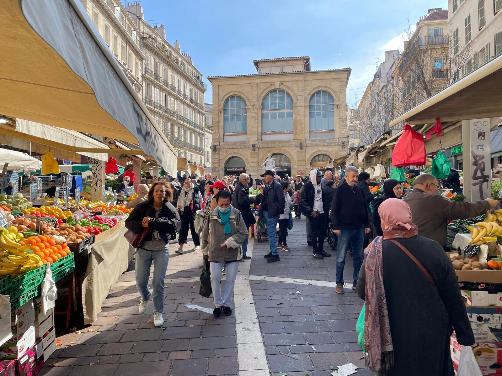 Fruit and vegetable stalls under awnings lining a narrow street at the Marché des Capucins in Marseille's Noailles quarter, crowded with shoppers, with an arcaded stone building closing the far end