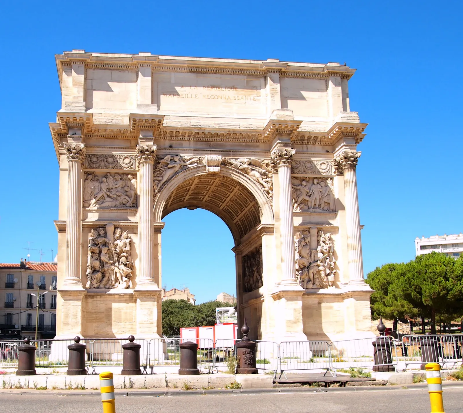 The Porte d'Aix triumphal arch in Marseille, a single pale stone arch with Corinthian columns and carved relief panels, standing alone on Place Jules-Guesde under a clear sky