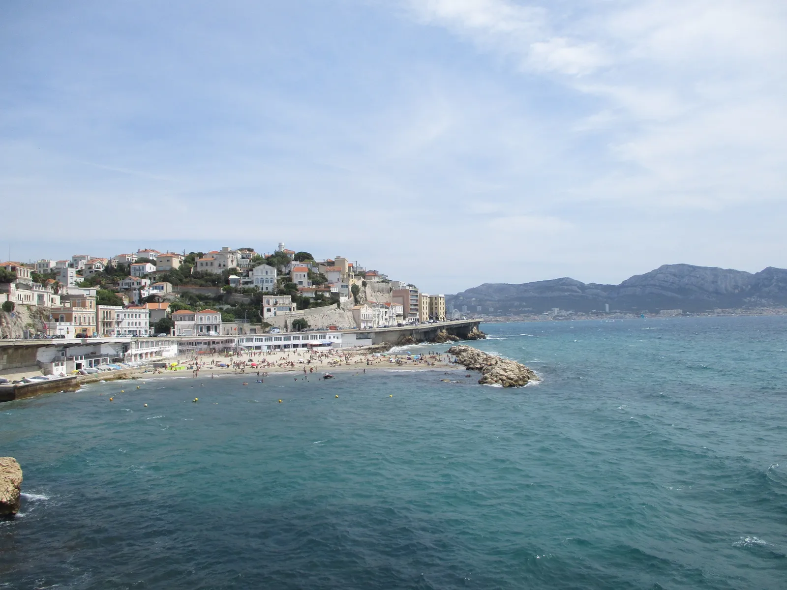 The coast along the Corniche Kennedy in Marseille, a small beach and swimmers below a hillside of pale villas, with the open sea and the hills of the Calanques beyond