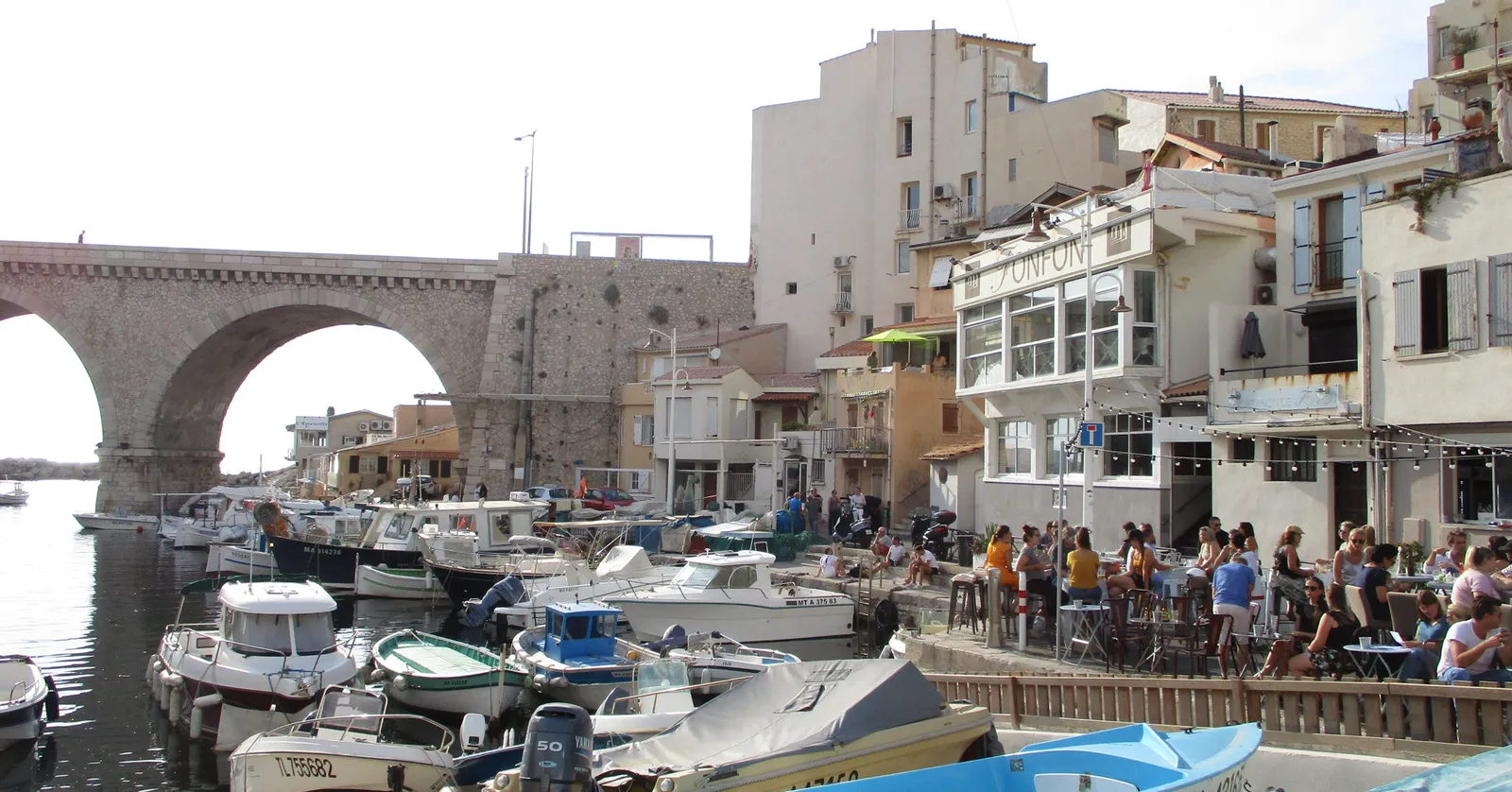 The fishing cove at Vallon des Auffes in Marseille, small boats moored along the quay under the stone arches of the road viaduct, with restaurant terraces full of people on the right
