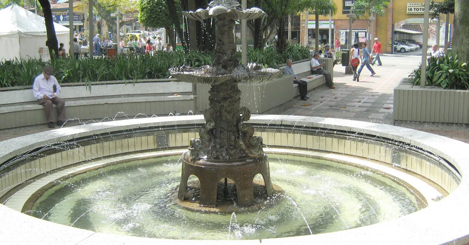 A tiered stone fountain in Parque Principal de Envigado, Medellín, with locals sitting on benches under trees around it