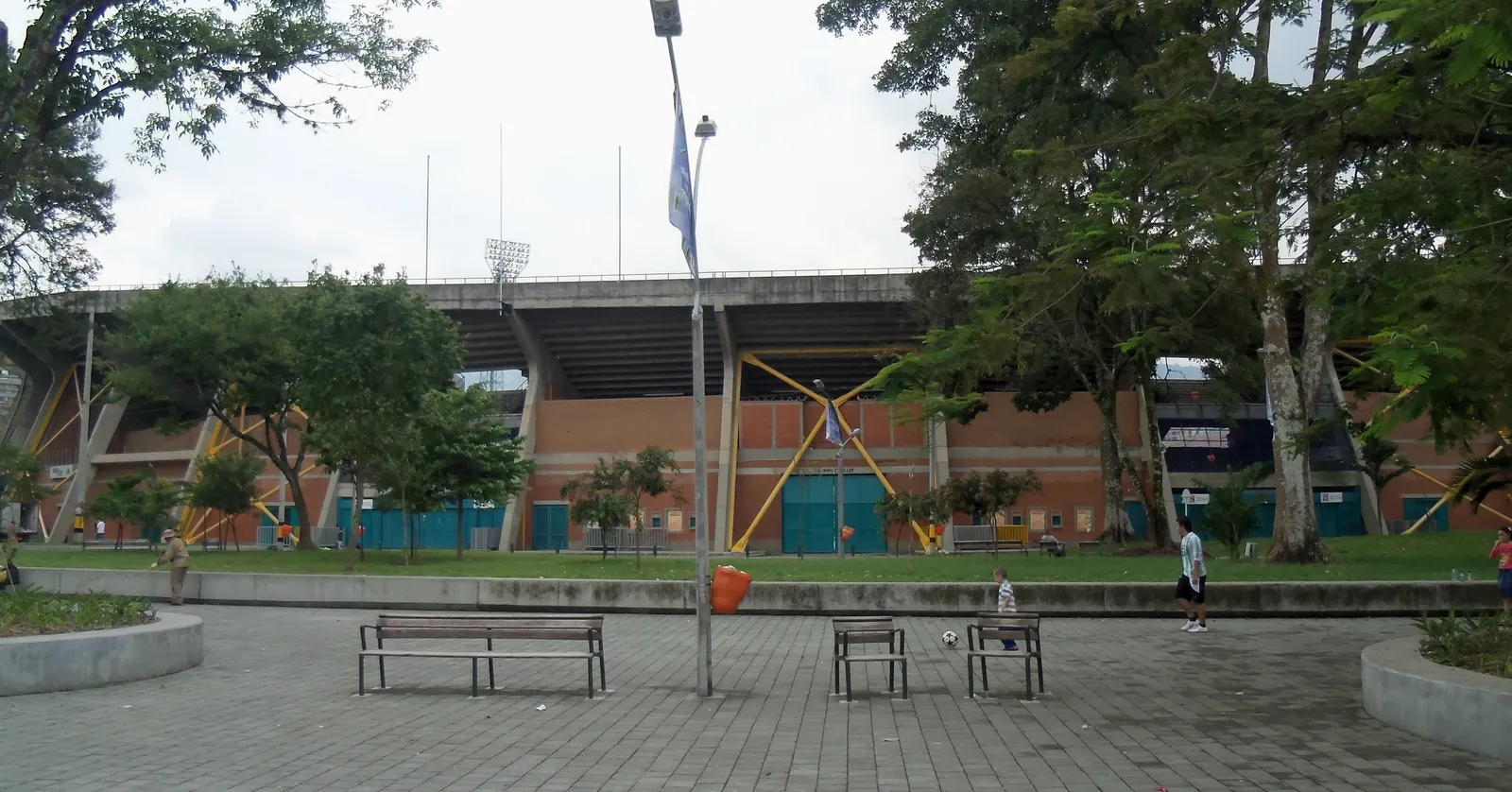 The brick and concrete exterior of Estadio Atanasio Girardot in Medellín, with people playing football on the grass in front of it