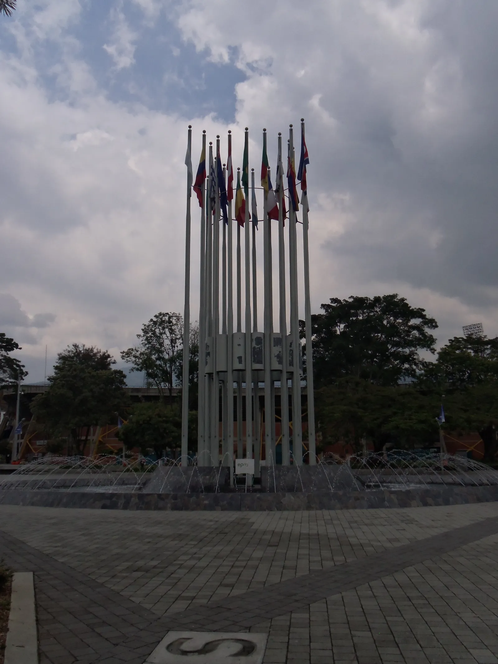 The flagpole monument and fountain at Plaza de Banderas inside Medellín's Atanasio Girardot sports complex, with international flags flying against a cloudy sky