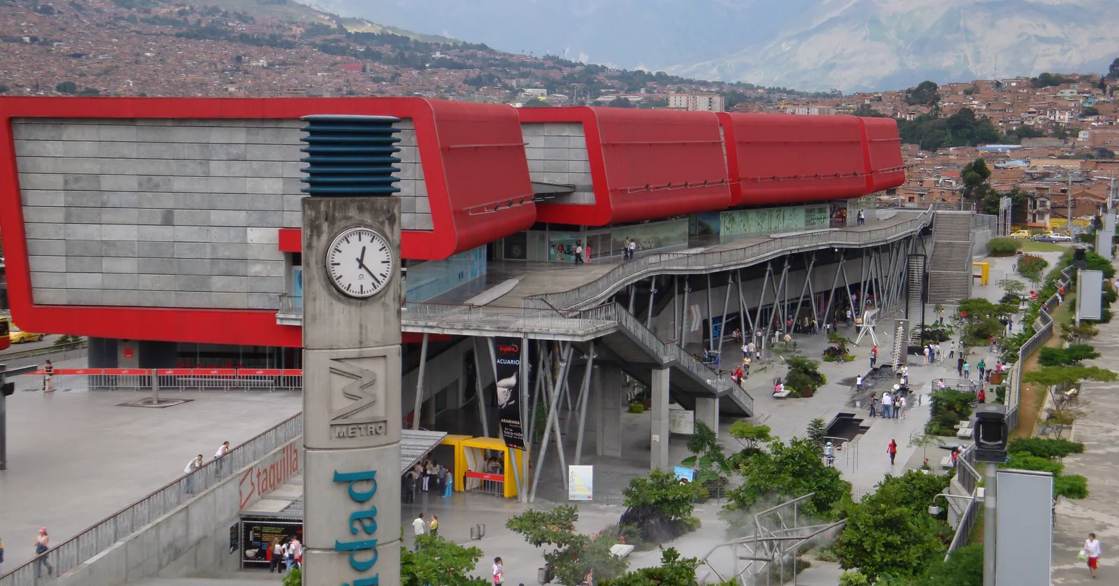 Parque Explora's red, wave-shaped roof in Medellín, seen from the elevated Universidad metro platform with the city's hillside neighborhoods behind it